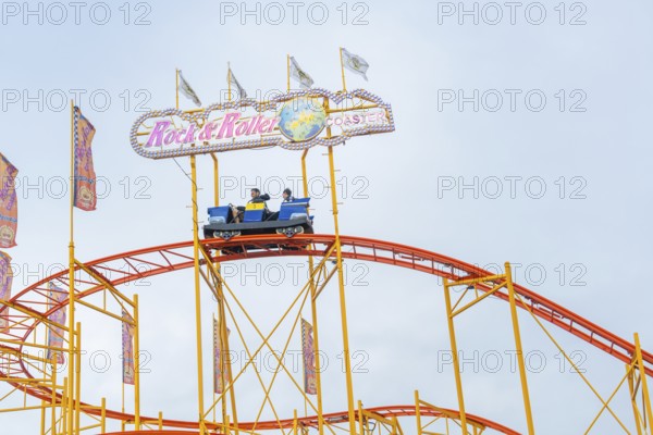 People in a roller coaster car on an orange-coloured track in front of colourful flags, Cannstatter Wasen funfair, Stuttgart, Germany