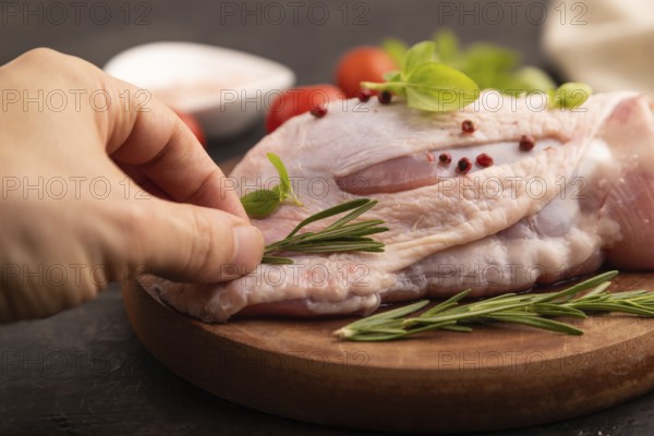 Raw Turkey Thigh with spices and rosemary on cutting board on black concrete background with hand and linen textile. side view, close up, selective focus