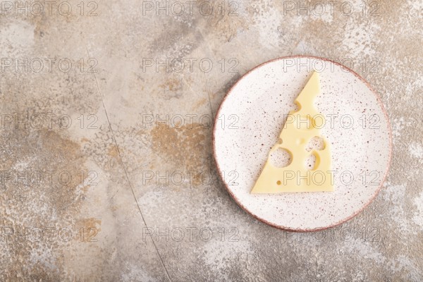 Piece of cheese with holes on ceramic plate on brown concrete background, top view, flat lay, copy space, minimalism
