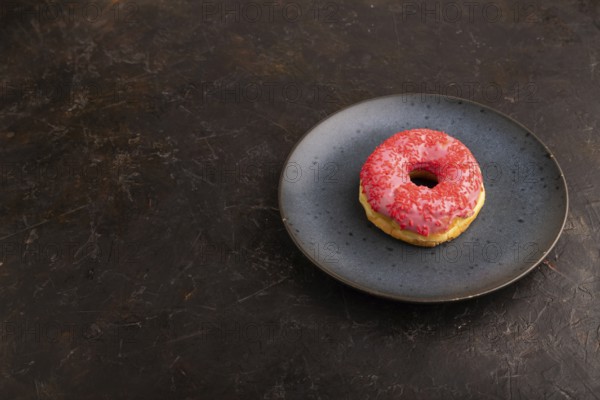 Pink Donut with sprinkles on blue ceramic plate on black concrete background, side view, copy space, minimalism