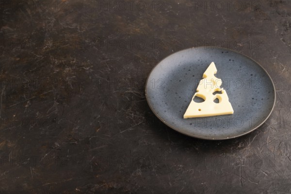 Piece of cheese with holes on blue plate on black concrete background, side view, copy space, minimalism