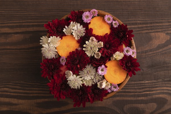 Wooden bowl with carrot slices and red Chrysanthemum flowers, Astrantia flowers, flower salad on brown wooden background, top view, flat lay, close up, minimalism
