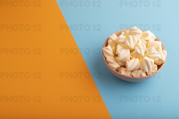 Orange and pink marshmallow in ceramic bowl on blue and orange pastel paper background, side view, copy space, minimalism
