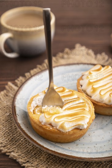 French lemon tart with meringue on brown wooden background, cup of coffee, linen textile, side view, close up, selective focus