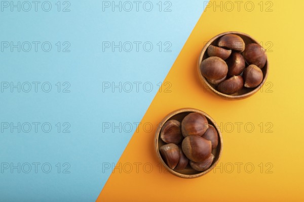 Wooden bowl with raw edible ?hestnuts on blue and orange paper pastel background, top view, flat lay, copy space, minimalism