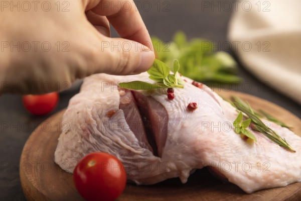 Raw Turkey Thigh with spices and rosemary on cutting board on black concrete background with hand and linen textile. side view, close up, selective focus