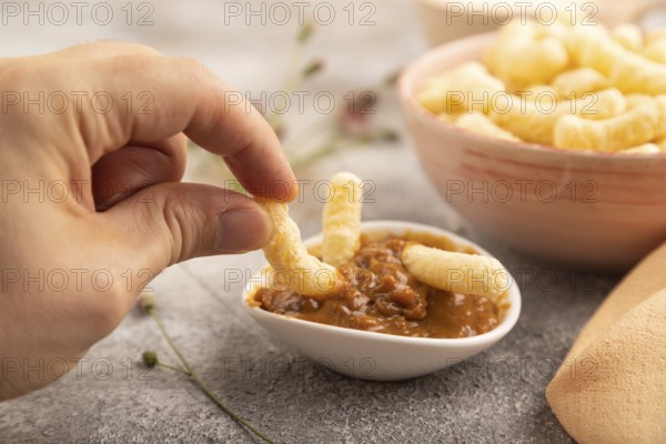 Corn flakes sticks with hand with caramel in ceramic bowl on gray concrete background and orange linen textile. Side view, close up