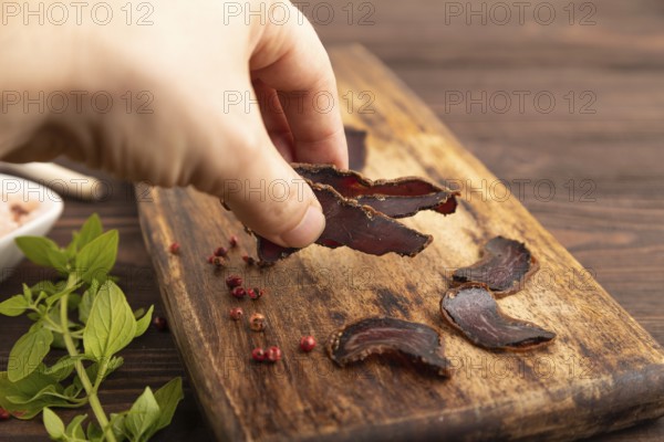 Armenian Basturma dried meat with hand on wooden cutting board with pepper and herbs on brown wooden background. Side view, close up, selective focus