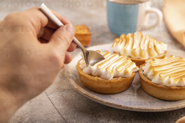 French lemon tart with meringue with hand on brown concrete background, cup of coffee, orange linen textile, side view, close up, selective focus
