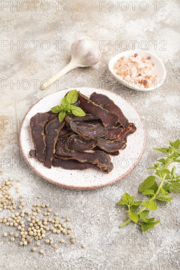 Armenian Basturma dried meat on plate with pepper and herbs on brown concrete background. Side view, close up