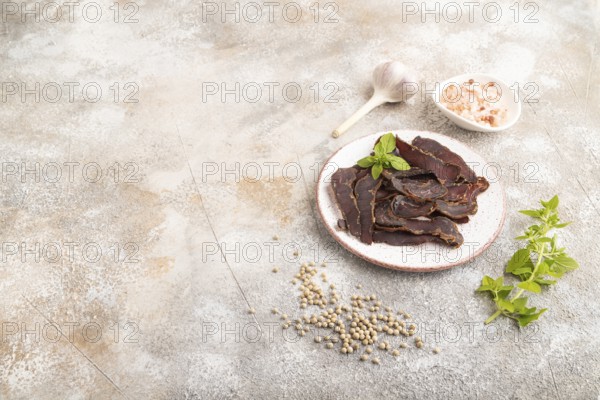 Armenian Basturma dried meat on plate with pepper and herbs on brown concrete background. Side view, copy space