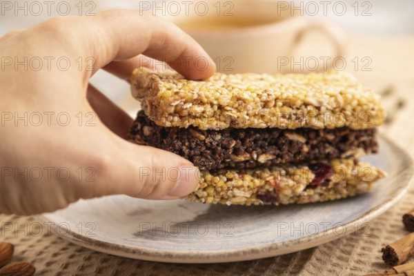 Granola bars with caramel, nuts, flakes in ceramic plate on brown concrete background with hand, beige linen napkin, cup of green tea. Side view, close up, selective focus