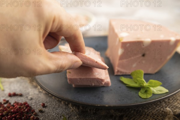 Mortadella sausage with hand on plate with pepper and herbs on brown concrete background and linen textile. Side view, close up, selective focus