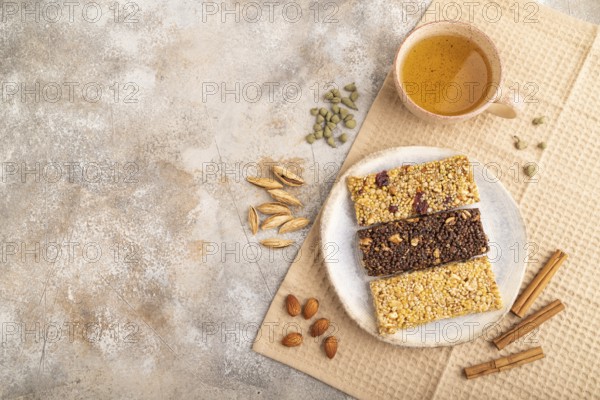 Granola bars with caramel, nuts, flakes in ceramic plate on brown concrete background, beige linen napkin, cup of green tea. Top view, flat lay, copy space