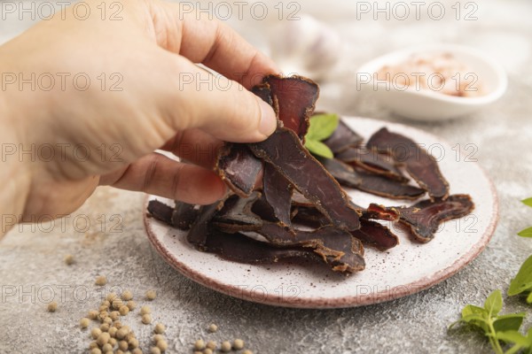 Armenian Basturma dried meat with hand on plate with pepper and herbs on brown concrete background. Side view, close up, selective focus