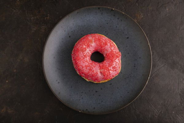 Pink Donut with sprinkles on blue ceramic plate on black concrete background, top view, flat lay, close up, minimalism