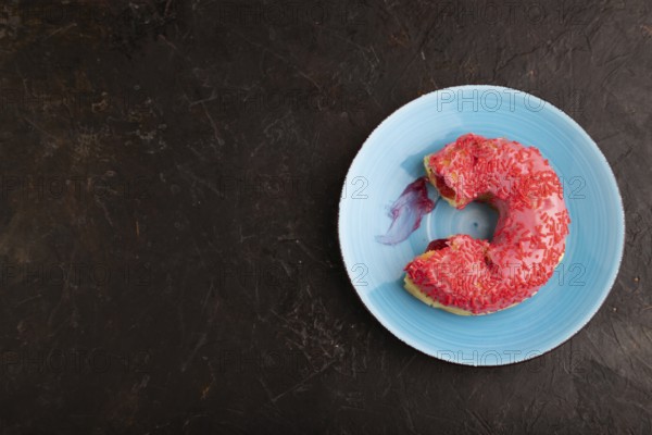 Bitten Pink Donut with sprinkles on blue ceramic plate on black concrete background, top view, flat lay, copy space, minimalism