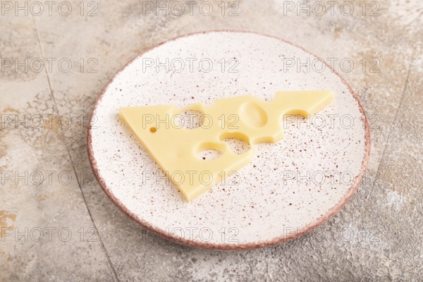 Piece of cheese with holes on ceramic plate on brown concrete background, side view, close up, minimalism