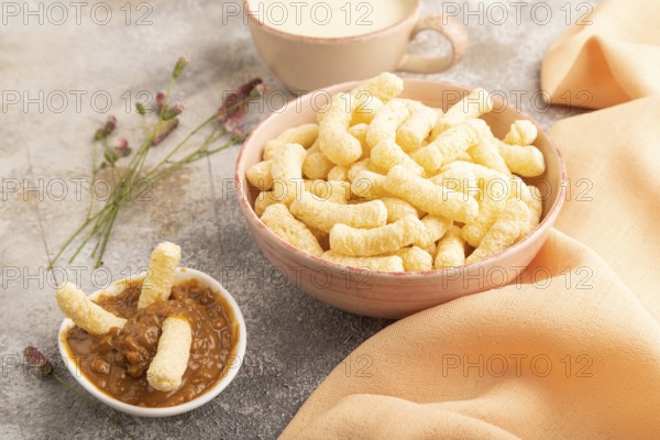 Corn flakes sticks with caramel in ceramic bowl on gray concrete background and orange linen textile. Side view, close up