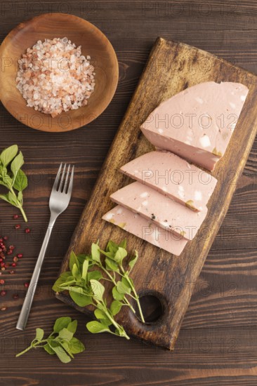 Mortadella sausage on wooden cutting board with pepper and herbs on brown wooden background. Top view, flat lay, close up