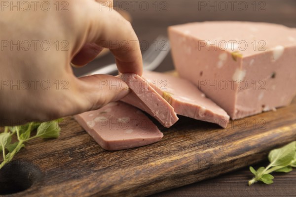 Mortadella sausage with hand on wooden cutting board with pepper and herbs on brown wooden background. Side view, close up, selective focus