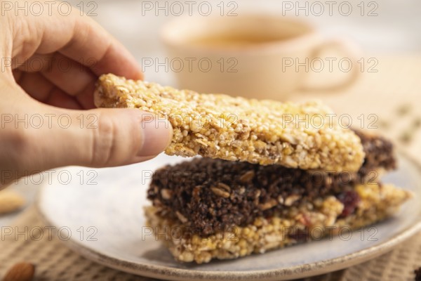 Granola bars with caramel, nuts, flakes in ceramic plate on brown concrete background with hand, beige linen napkin, cup of green tea. Side view, close up, selective focus