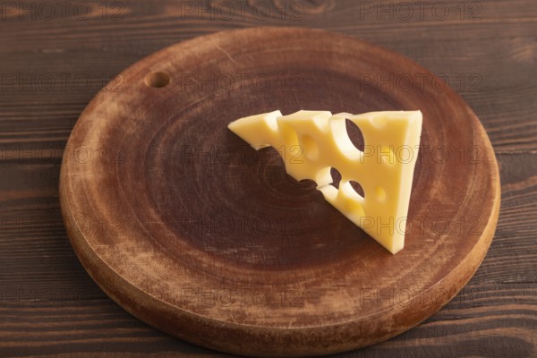 Piece of cheese with holes on cutting board on brown wooden background, side view, close up, minimalism