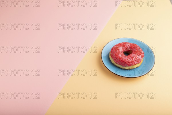Pink Donut with sprinkles on blue ceramic plate on blue and orange pastel paper background, side view, copy space, minimalism