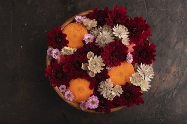 Wooden bowl with carrot slices and red Chrysanthemum flowers, Astrantia flowers, flower salad on black concrete background, top view, flat lay, minimalism, close up