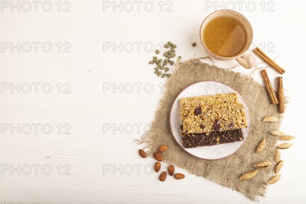 Granola bars with caramel, nuts, flakes in ceramic plate on white wooden background, beige linen napkin, cup of green tea. Top view, flat lay, copy space