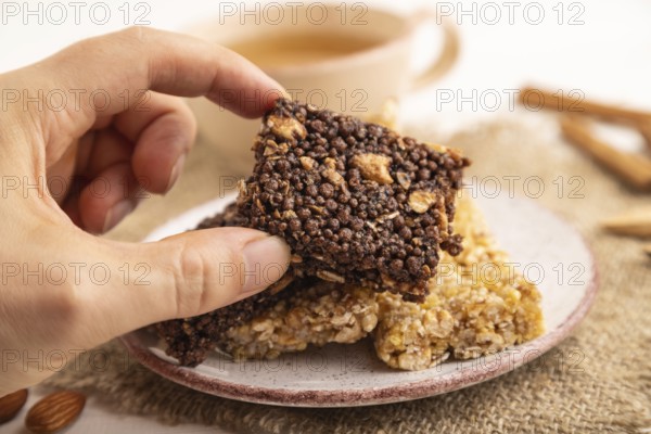 Granola bars with caramel, nuts, flakes in ceramic plate with hand on white wooden background, beige linen napkin, cup of green tea. Side view, close up, selective focus