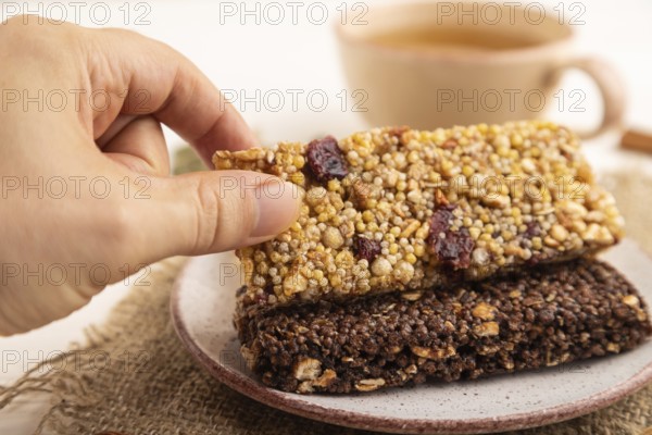 Granola bars with caramel, nuts, flakes in ceramic plate with hand on white wooden background, beige linen napkin, cup of green tea. Side view, close up, selective focus