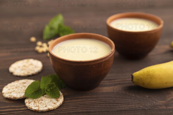 Organic non dairy banana and soy milk in clay cup on brown wooden background. Vegan healthy food concept, side view, close up, selective focus