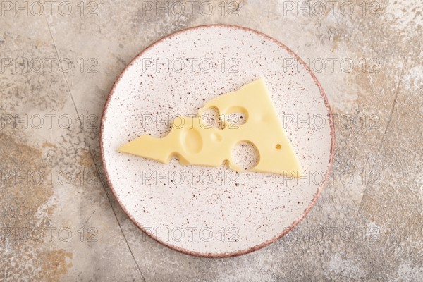 Piece of cheese with holes on ceramic plate on brown concrete background, top view, flat lay, copy space, minimalism