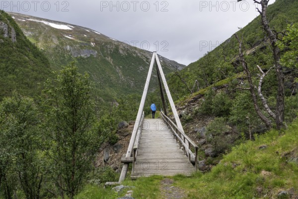 Hiker with rucksack and blue raincoat crosses wooden bridge, Kongevegen or Kingsroad, pilgrimage route Olavsweg or Olavsleden, Drivdalen valley, ascent to Dovrefjell, Oppdal municipality, Trøndelag county, Norway