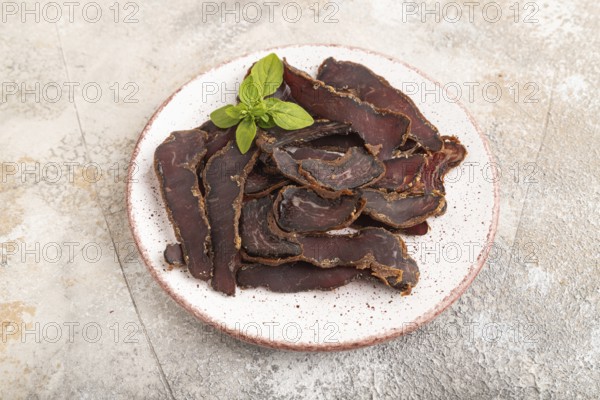 Armenian Basturma dried meat on plate with pepper and herbs on brown concrete background. Side view, close up