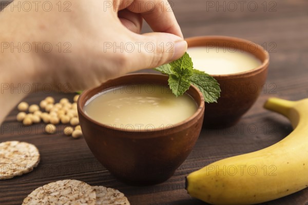 Organic non dairy banana and soy milk in clay cup with hand on brown wooden background. Vegan healthy food concept, side view, close up, selective focus