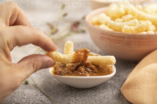 Corn flakes sticks with hand with caramel in ceramic bowl on gray concrete background and orange linen textile. Side view, close up