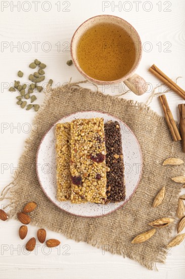 Granola bars with caramel, nuts, flakes in ceramic plate on white wooden background, beige linen napkin, cup of green tea. Top view, flat lay, close up