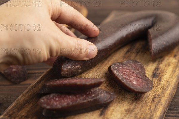 Sujuk sausage with hand on wooden cutting board with pepper and herbs on brown wooden background. Side view, close up, selective focus