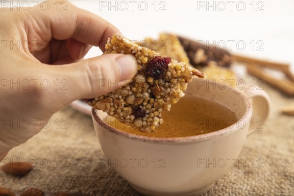 Granola bars with caramel, nuts, flakes in ceramic plate with hand on white wooden background, beige linen napkin, cup of green tea. Side view, close up, selective focus