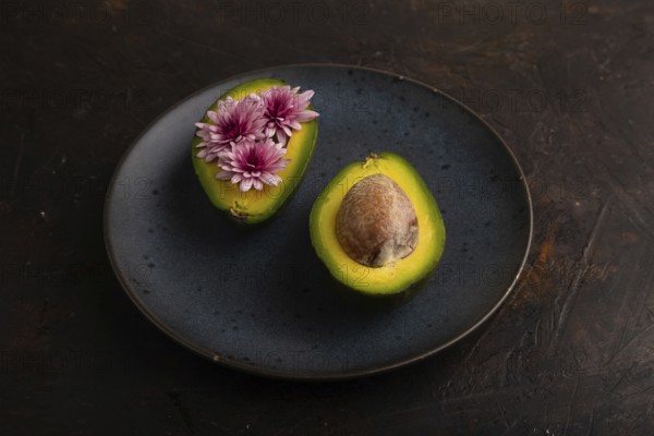 Two Avocado halves with pit and Chrysanthemums on blue plate on black concrete background, side view, minimalism, low key, close up