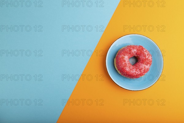 Pink Donut with sprinkles on blue ceramic plate on blue and orange pastel paper background, top view, flat lay, copy space, minimalism