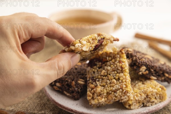 Granola bars with caramel, nuts, flakes in ceramic plate with hand on white wooden background, beige linen napkin, cup of green tea. Side view, close up, selective focus