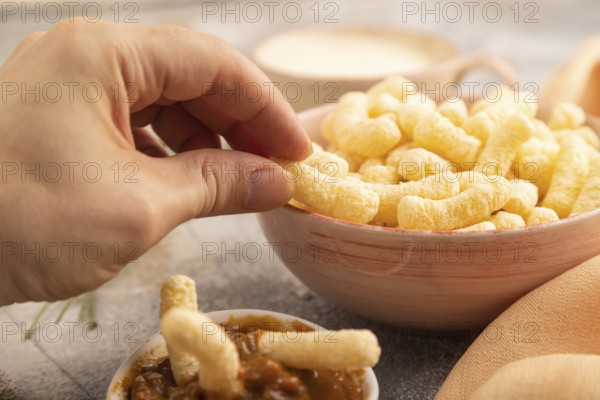Corn flakes sticks with hand with caramel in ceramic bowl on gray concrete background and orange linen textile. Side view, close up