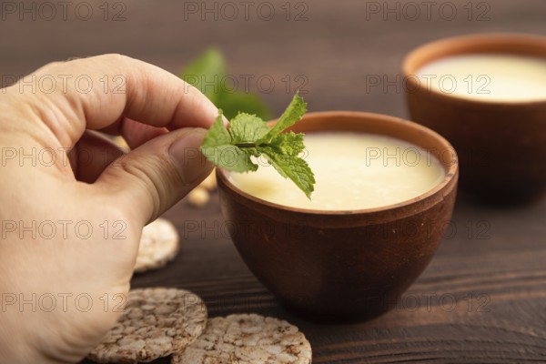Organic non dairy banana and soy milk in clay cup with hand on brown wooden background. Vegan healthy food concept, side view, close up, selective focus