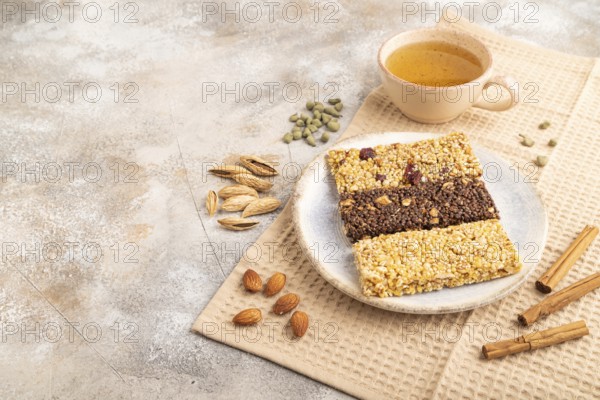 Granola bars with caramel, nuts, flakes in ceramic plate on brown concrete background, beige linen napkin, cup of green tea. Side view, copy space