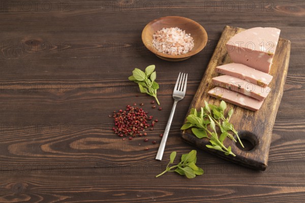 Mortadella sausage on wooden cutting board with pepper and herbs on brown wooden background. Side view, copy space