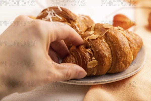 Croissant on blue plate with hand on white wooden background and orange linen textile, side view, close up, selective focus