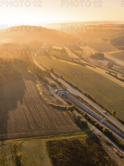 Landschaft während eines Sonnenaufgangs mit Feldern, Hügeln und einer Straße, die von goldenem Licht beleuchtet wird, Neubautunnel der Hermann Hessebahn, Calw, Schwarzwald, Deutschland